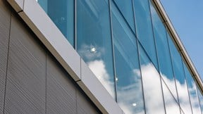 Modern glass office façade reflecting clouds against a clear blue sky, highlighting the building's clean lines and contemporary design.