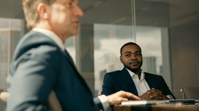 Two people are seated at a meeting table in a modern office conference room. One person is in sharp focus on the right side of the image, wearing a dark blazer and white collared shirt, seated with hands clasped on the table. Another person is visible in the foreground on the left, softly out of focus, creating depth in the composition. A notebook and an open laptop rest on the table, while glass walls and reflections of the office interior form the background. Natural daylight illuminates the space, and a shallow depth of field keeps the person on the right in focus while the foreground and background remain softly blurred.