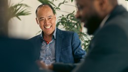 A professional seated in a meeting room, wearing a blue blazer and striped shirt, viewed between two blurred colleagues in the foreground. Indoor plants
