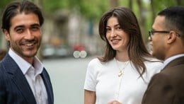 Three people are outdoors, engaged in conversation. They appear happy and relaxed, with blurred greenery in the background.