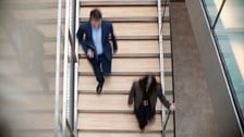 A blurred overhead shot of two people walking on a wooden staircase in a modern building. The movement conveys a sense of urgency and busy office life.