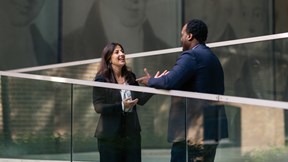Two colleagues standing outside New Court, speaking beside a glass railing with glass reflections of the five brothers visible in the background.