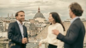Three people stand on a rooftop overlooking a cityscape. The atmosphere is lively, with one person gesturing animatedly while chatting.