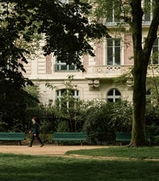 Views of classic Paris architecture between the trees of a park, with a person walking in the foreground